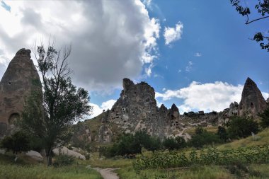 Kapadokya 'nın alışılmadık manzarası. Antik Uchisar kalesi mavi gökyüzünün arka planına karşı yükselir. Birçok mağara kayalara oyuldu. Yol yeşil bir çayırdan geçer ve ağaçların arasından