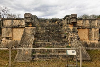 Antik Maya şehri Chichen Itza 'nın mimarisi. Basamaklar taş platformun tepesine çıkıyor. Yan tarafında, taştan oyulmuş bir yılanın kafası var. Gökyüzü bulutlu. Meksika
