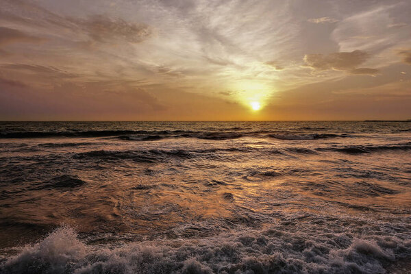Золотой закат над Индийским океаном. В небе светлые перистые облака. Волны катятся к берегу и пена на пляже. Шри-Ланка. Коломбо.