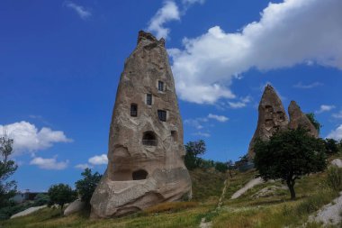 Mavi gökyüzünün arka planında, bir tepede konik bir kaya yükselir. Modern pencereler antik mağaralara yerleştirilmiştir. Patika çayırdan geçerek uzaklığa gidiyor. Kapadokya. Türkiye