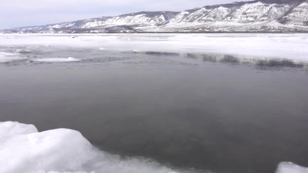 氷に覆われた湖の雪に囲まれて帽をかぶった山々 解凍されたパッチ内の水が風から揺れています 氷の一片が縁から切り出されて浮く ロシアだ バイカル — ストック動画
