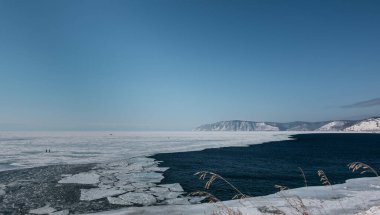 Angara, Baykal Gölü 'nden buzla kaplı olarak çıkar. Nehrin ağzı donmaz, buz kütleleri mavi suda yüzer. Ön planda kuru çimen çıkıntıları var. Uzakta dağlar.