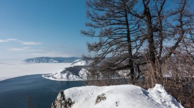 Baykal Gölü 'nün buzla kaplı üst manzarası ve Angara Nehri' nin donmayan kaynağı. Ön planda çıplak ağaçlar ve bir kaya var. Uzakta, karla kaplı dağlar.