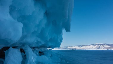 Kayanın tabanı kalın bir mavi buz tabakası ve sıçramalarla kaplı. Gölün yüzeyinde kar var. Uzakta bir dağ sırası görülebilir. Baykal Gölü. Rusya