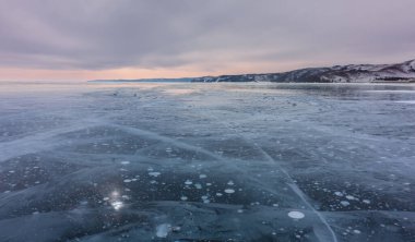 Baykal Gölü üzerinde kış günbatımı. Buzun pürüzsüz yüzeyi çatlaklarla kaplı. Donmuş metan gazından oluşan beyaz kabarcıklar görülebilir. Akşam gökyüzünün arka planında dağların siluetleri.