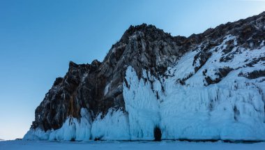 Kayanın dik yamaçları tuhaf buz parçacıkları ve sarkıtlarla kaplıdır. Üste küçük koyu bir mağara var. Donmuş bir gölün yüzeyinde kar. Mavi gökyüzü. Baykal.