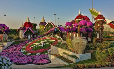 Parkta saat şeklinde, elleri ve tuşları olan renkli bir çiçek tarhı var. Etrafta bir sürü parlak renkli çiçek aranjmanı var..