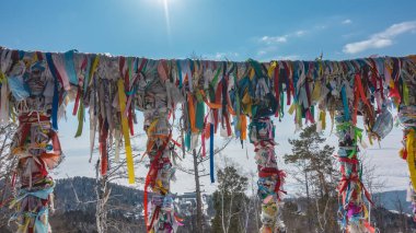 Parlak renkli kurdeleler ve tek kullanımlık tıbbi maskeler gözlem güvertesinin korkuluklarına bağlıdır. Şamanizm ayini. Salgının özellikleri. Arkaplan - mavi gökyüzü, donmuş Baykal Gölü.