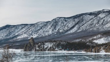 Donmuş göl üzerinde dik kayalık yamaçları olan görkemli bir uçurum. Buzda yansıma. Kıyıda küçük bir köy var. Arka planda karla kaplı ağaçlık dağlar var. Baykal.