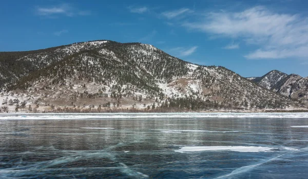 凍った湖の氷の滑らかな光沢のある表面には 雪の亀裂や領域が見えます 海岸では 青空の背景には 木製の山があります バイカル — ストック写真