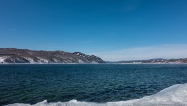 Kışın Angara nehri. Mavi suda dalgalanmalar. Kıyıda kar ve buz var. Gök kubbesinin arkaplanına karşı sıradağlar.