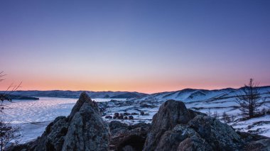 Kış gündoğumu. Dağların üstündeki gökyüzü pembeyle işaretlenmiş. Donmuş bir gölün buzunda bir güneş parıltısı. Vadideki köy evleri. Ön planda pürüzlü bir dokusu olan kayalar var. Baykal.