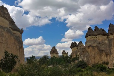 Kümülüs bulutlu mavi gökyüzüne karşı yüksek inanılmaz kayalar. Sivri tepeli silindirik uçurumlar, mantar kapaklarına benzer, sıra halinde dururlar. Çevrede yeşil bitki örtüsü. Kapadokya. Türkiye