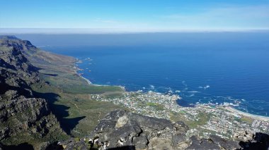 Güney Afrika kıyı şeridi yukarıdan. Mavi Atlantik Okyanusu, dalgaların köpüğü, gökyüzü, düzensiz kıyılar, şehir evleri. Ön planda Masa Dağı zirvesinin gri taşları var. Cape Town.