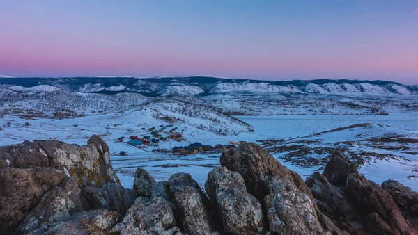 Dawn over a snowy valley. The sky above the ridge turns pink. Village houses on the shore of a frozen lake. In the foreground there are picturesque rocks. Stone texture. Baikal