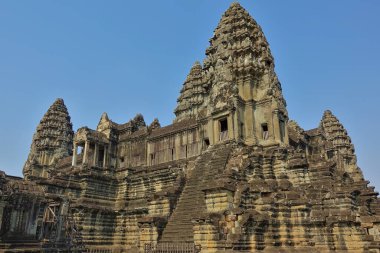 Angkor 'un antik tapınağı mavi gökyüzüne karşı. Yüksek kuleler oymalar ve kabartmalarla süslenir. Taş bir merdiven üst kattaki sütunlu terasa çıkıyor. Kamboçya.