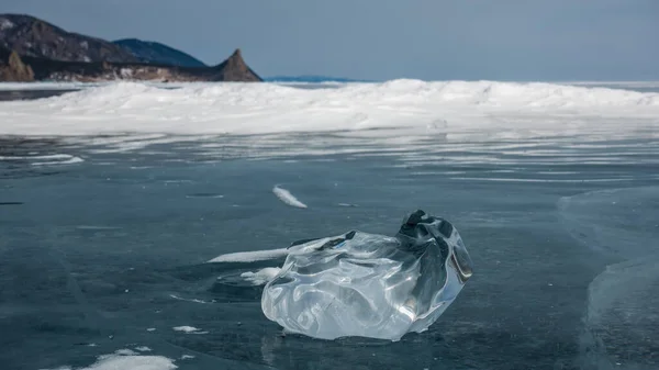 A shiny transparent shard of ice of a bizarre shape lies on the surface of a frozen lake. Rounded, curved edges. Reflection. In the distance there are snowdrifts, a mountain range. Baikal