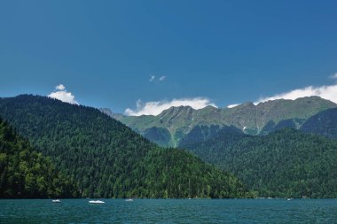Beyaz kayıklar turkuaz alp gölü Ritsa 'da yüzer. Kıyılarda kozalaklı ormanlarla kaplı dağlar var. Parlak mavi gökyüzünde kümülüs bulutları var. Abhazya