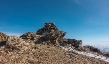 Bitkilerden yoksun, açık mavi bir gökyüzünün arkaplanına bakan resimli kayalıklar. Taşlarda çatlaklar var. Zeminde biraz kar var. Donmuş gölün yüzeyi görülebilir. Baykal.