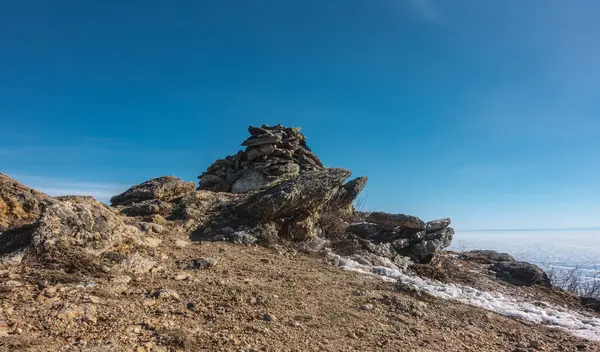 Bitkilerden yoksun, açık mavi bir gökyüzünün arkaplanına bakan resimli kayalıklar. Taşlarda çatlaklar var. Zeminde biraz kar var. Donmuş gölün yüzeyi görülebilir. Baykal.