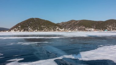 Donmuş gölün pürüzsüz yüzeyinde kar parçaları görülebilir. Derin çatlaklar şeffaf mavi buzu geçer. Kıyıda ağaçlık dağlar ve bir köy var. Gök mavisi. Kış günü. Baykal.