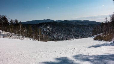 Bir teleferik karla kaplı dağ yamacının üzerinden geçer. Karda kayak izleri. Berrak bir gökyüzünün arka planında ağaçlandırılmış dağ sıraları. Kış günü
