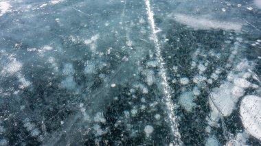 Turkuaz şeffaf buz, yakın plan, tam kare. Yüzeyde çatlaklar ve paten izleri var. Derinliklerde donmuş metan gazı baloncukları görünür. Baykal Gölü.