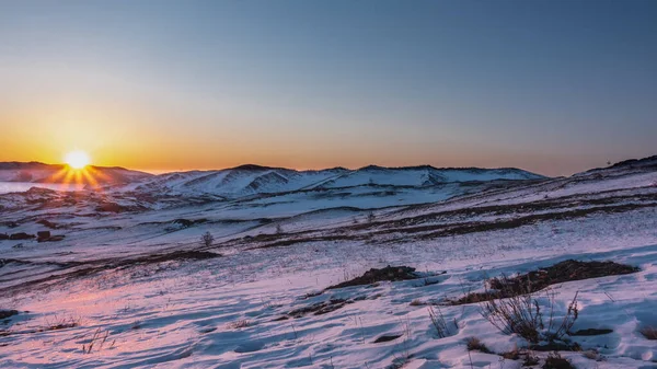 Sunrise over a snowy plain. The sun is over the ridge, its rays color the snow in golden hues. Dry grass and rare shrubs on the ground. The frozen Lake Baikal is visible.