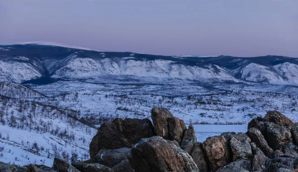 Early morning. The snow-covered valley is surrounded by a mountain range. The sky is turning pink. In the foreground there are picturesque granite rocks devoid of vegetation. Siberia