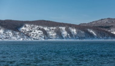 Donmayan Angara nehri. Mavi suda dalgalanmalar. Kıyıda, karla kaplı bir dağın yamacında köy evleri ve orman görülebilir. Açık gökyüzü, güneşli kış günü.