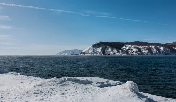 stock image The non-freezing Angara river in winter. The banks are covered with snow. Ripples in the blue water. Snow-covered wooded mountain range against the backdrop of a clear sky.