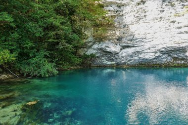 Göl dik kayalıklarla çevrilidir. Aquamarine suyu sakin ve temiz. Yansıma. Taşlar ve algler görülebilir. Mavi Göl. Abhazya.