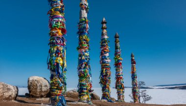 Uzun tahta sütunlar çok renkli ritüellerle bağlanır ve sıraya dizilir. Yakınlarda pitoresk kayalar var. Arkaplan - açık mavi gökyüzü, donmuş göl. Baykal. Şamanka Dağı