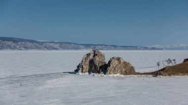 İki başlı, bitkisiz bir kaya donmuş gölde yükselir. Buzdaki küçük insan siluetleri. Karla kaplı dağ sıraları gökyüzüne karşı. Baykal.