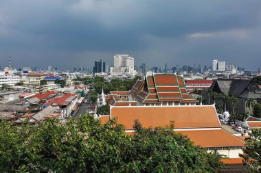 Bangkok şehir merkezi. Eski, modern, yeni ve yıpranmış binalar. Bulutlu gökyüzüne karşı gökdelenler. Ön planda yeşil ağaç dalları var. Tayland