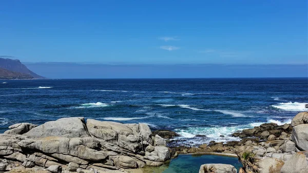 Atlantic coastline in Cape Town. There are picturesque boulders on the shore. There is foam on the blue waves. Mountain range against the background of the azure sky. South Africa