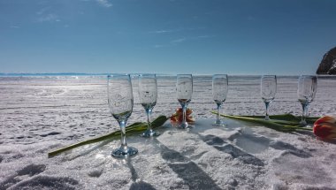 Tatil için hazırlık. Temiz boş şarap kadehleri karda dizilmiştir. Yakınlarda parlak kırmızı laleler var. Arka plan donmuş bir göl, açık mavi bir gökyüzü. Baykal.