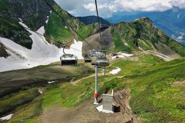 Yazın Kafkasya Dağları 'nda teleferik. Koltuklar boş. Dağların dik yamaçlarında kar var. Vadide yeşil çimenler var. Gökyüzünde resim gibi bulutlar. Rusya.
