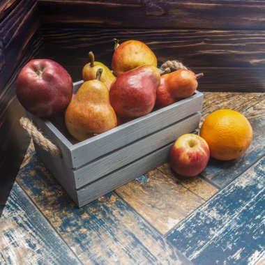 İp saplı mavi ahşap bir kutu olgun kırmızı armutlarla doludur. Yakınlarda, boyanmış tahtaların üzerinde bir elma ve bir portakal yatıyor. Yakın plan.