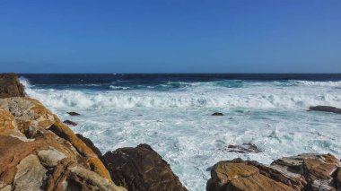Umudun Burnu açıklarındaki turkuaz Atlantik Okyanusu. Dalgalar kıyıya vurarak kalın bir köpük oluşturdu. Ön planda pitoresk kayalar var. Açık mavi gökyüzü. Güney Afrika