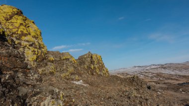Mavi gökyüzünün arka planında resimli bir dağ sırası. Sarı zambaklar dik granit yamaçlarda yetişir. Taşlarda çatlaklar var. Yerde kar var. Olkhon Adası. Baykal.