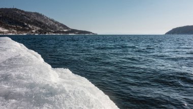 Kışın donmayan nehir. Mavi suda dalgalanmalar. Ön planda kıyıda bir kar yağışı var. Açık bir gökyüzünün arka planında karla kaplı dağlar. Sibirya 'da. Angara.