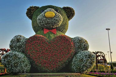 Bu büyük ayı yavrusu figürü yeşil bitkiler ve çiçeklerden yapılmıştır. Pençelerinde kırmızı petunyadan bir kalp taşıyor. Arka plan açık mavi gökyüzü. Dubai