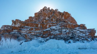 Mavi gökyüzüne karşı bitki örtüsünden yoksun kırmızı-kahverengi bir kaya. Garip şekiller, kayalardaki çatlaklar. Tabanda kalın bir buz saçağı tabakası var. Baykal.