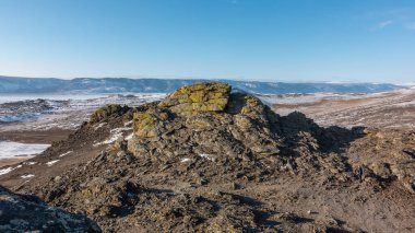 Kış Sibirya manzarası. Kırık granit kayaların üzerinde sarı lichenler yetişir. Uzakta, bir dağ sırası görebilirsiniz, donmuş bir göl. Açık mavi gökyüzü. Baykal.