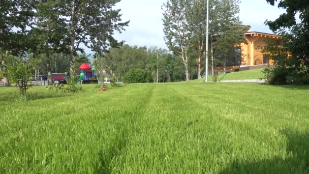 绿草草坪用自动灌溉系统浇水 水射流被喷出的机制和旋转 远处看得见孩子们的游乐场 阳光明媚的夏日 — 图库视频影像