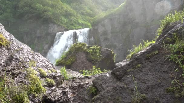 滝の力強い流れが崖の端から注ぐ 水滴が空中を飛ぶ 風が草を揺らす 前景に 絵のようにぬれた岩 カムチャツカ — ストック動画