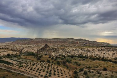 Kapadokya 'nın şafak vakti manzarası. Balonlar kayaların üzerinden katlanmış yamaçlar ve kırsal alanlarla uçar. Gökyüzünde gri bir bulut var. Uzakta yağmur yağıyor..