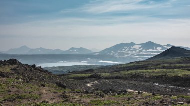 Dağ yamacında çok az bitki örtüsü var, volkanik kayalar. Uzakta bir göl görebilirsiniz. Mavi gökyüzüne karşı karla kaplı bir dağ sırası. Kamçatka.