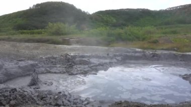 Doğal kaplıcalar. Yerdeki deliklerde gri sıvı kaynıyor. Sprey etrafta uçuşuyor. Buhar yükseliyor. Toprakta sülfür birikintileri. Yakınlarda yeşil bir tepe var. Kamçatka.